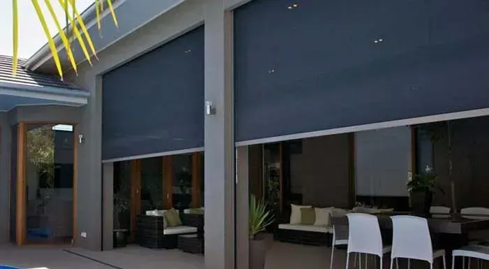 Dark gray outdoor roller blinds partially lowered over a patio with furniture visible inside.