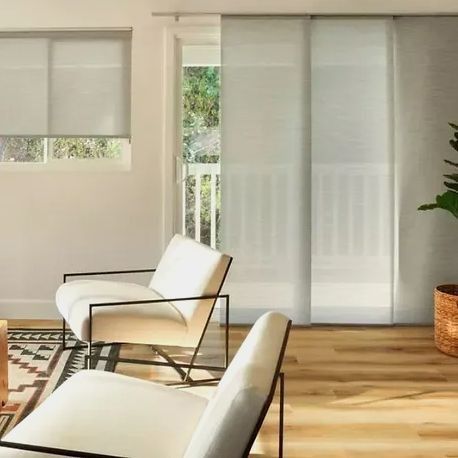 Living room with light-colored furniture, hardwood floors, and sheer panel shades. A balcony is visible through the door.