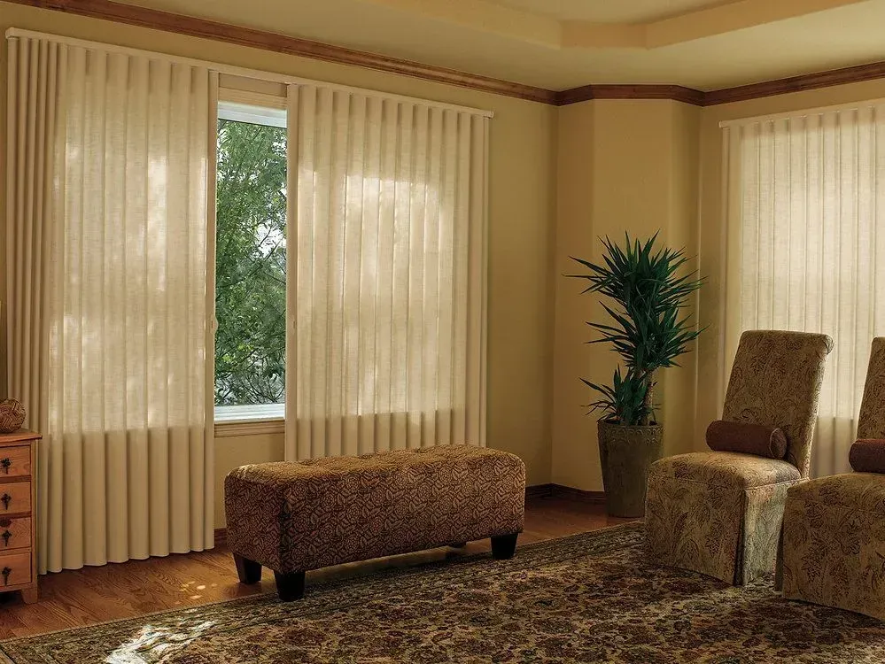 Living room with vertical blinds, beige walls, seating, and a window overlooking greenery.