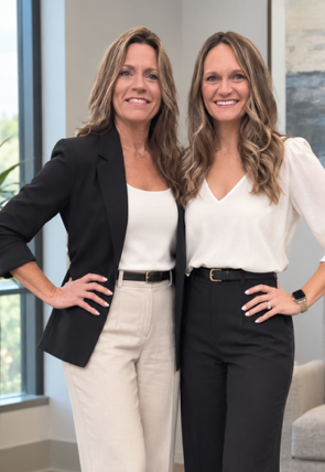 Two women standing indoors, smiling, in black-and-white outfits with hands on hips.