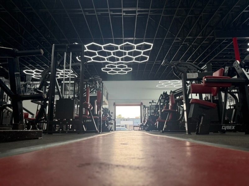 A gym interior with exercise machines, overhead honeycomb lights, and a red walkway.