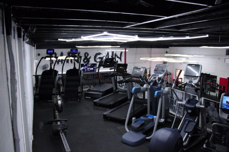 Gym interior with cardio machines: treadmills, ellipticals, stair steppers, and rowers. Black floor and ceiling, white walls.
