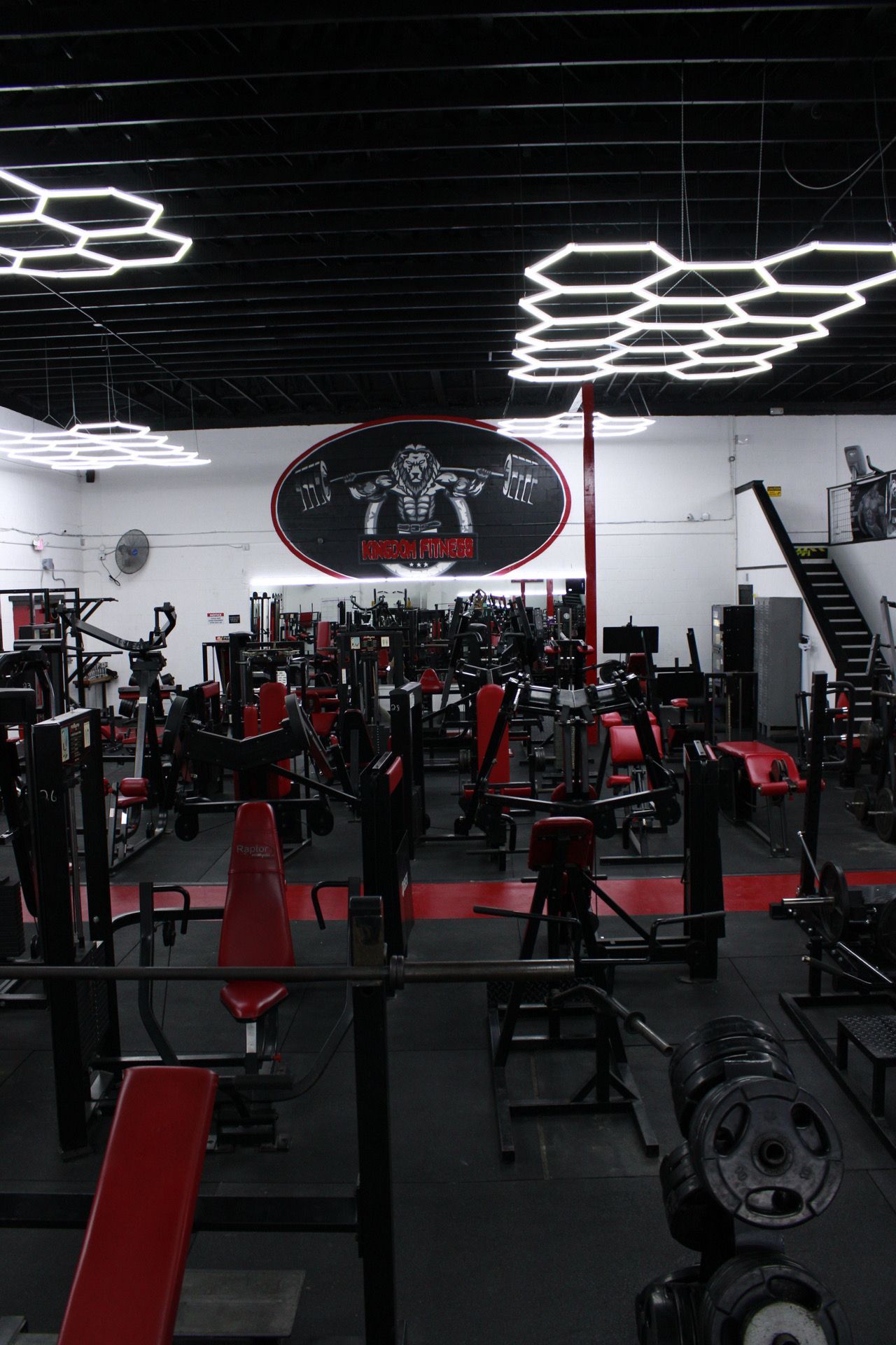Weightlifting gym with red and black equipment, logo on the back wall, and hexagonal ceiling lights.