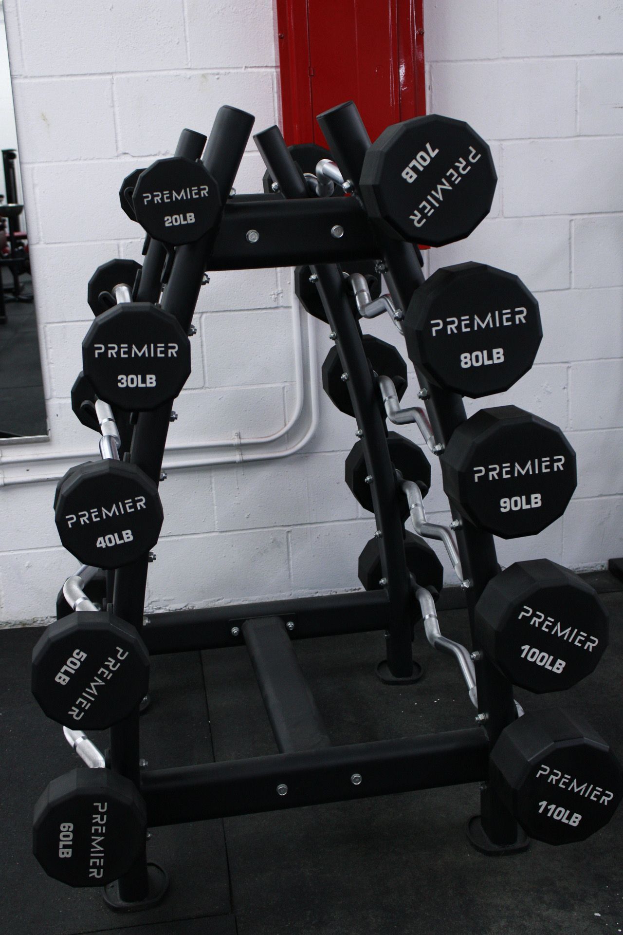 A rack holding several black dumbbells with weights ranging from 30 to 115 lbs. In a gym setting.