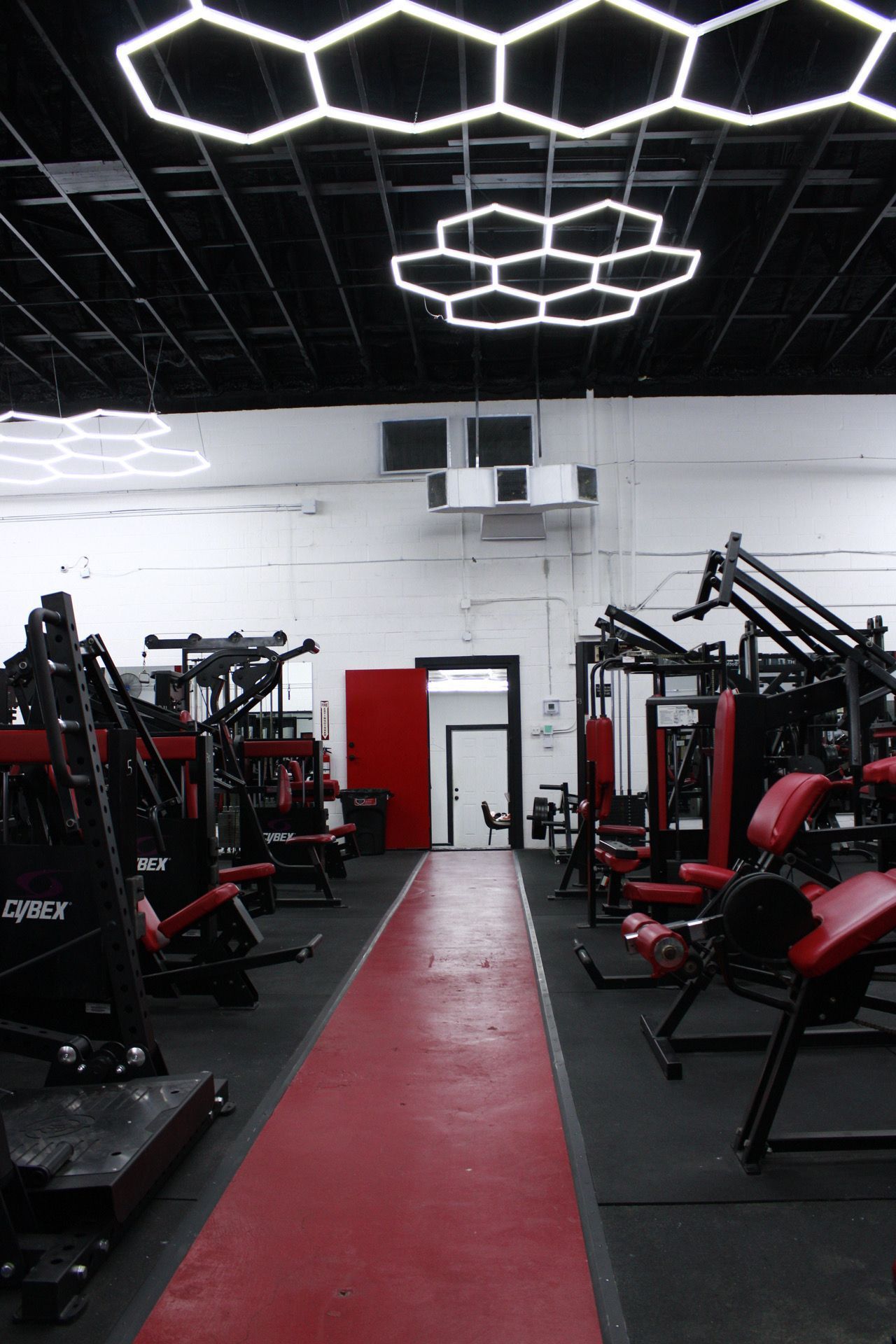 Gym interior with rows of exercise machines; red and black color scheme. Hexagonal lights on the ceiling.