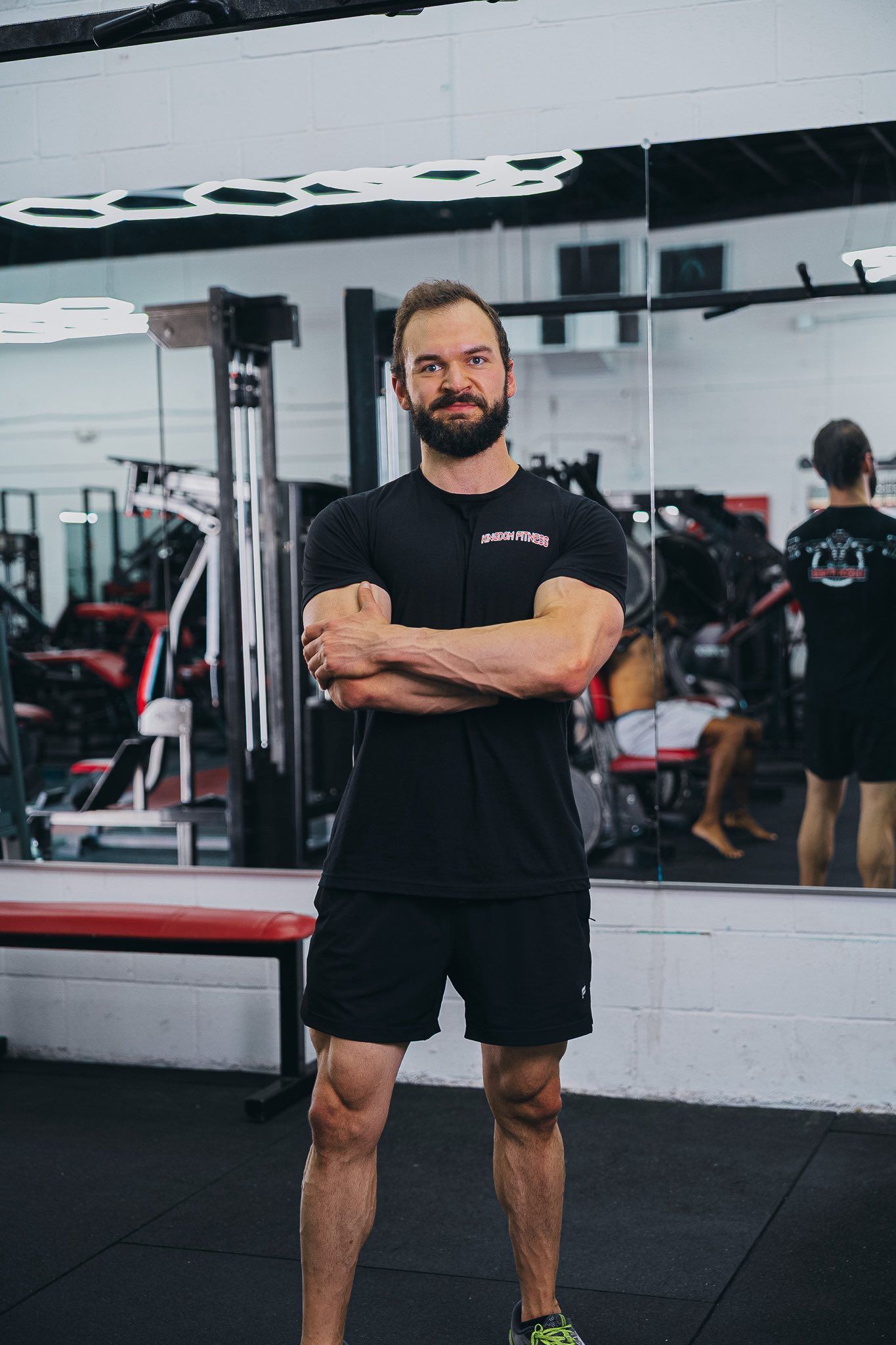 Man in gym with arms crossed, wearing black t-shirt and shorts. Standing in front of a mirror, gym equipment in background.