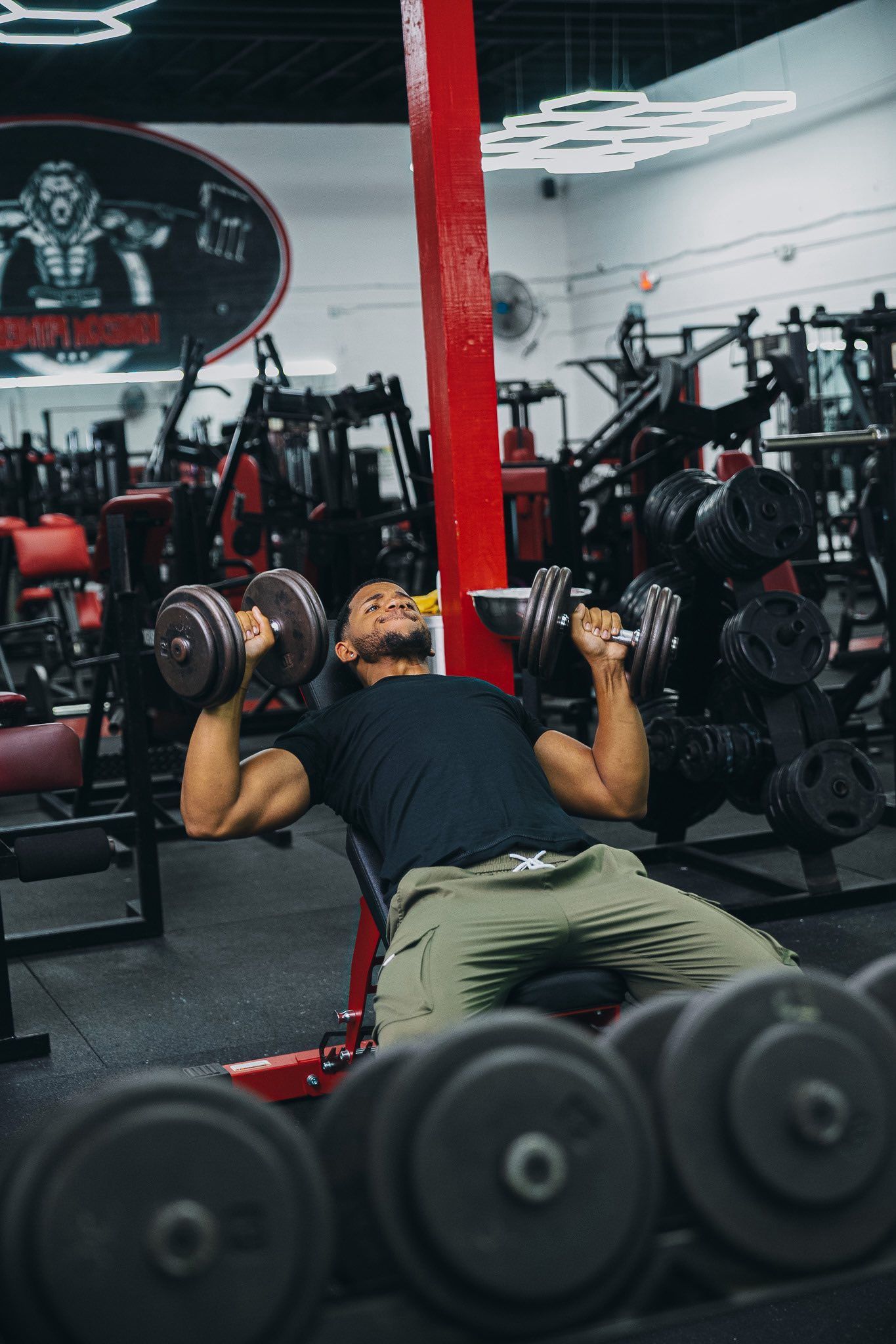 Man lifting dumbbells on a bench in a gym. Red and black color scheme.