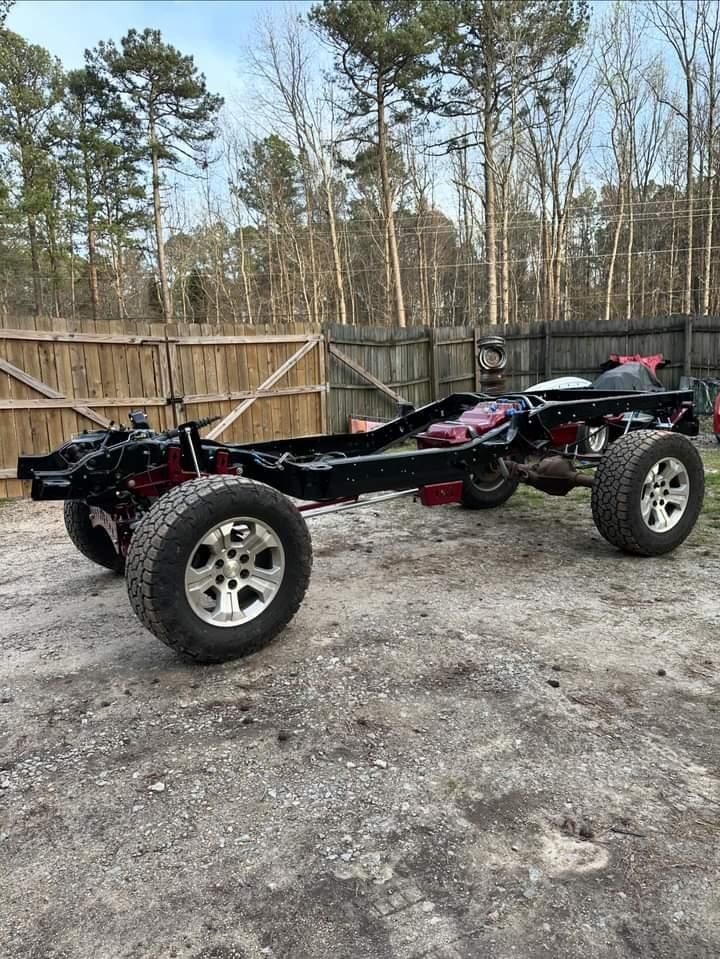 A jeep chassis is sitting on top of a dirt road next to a wooden fence.