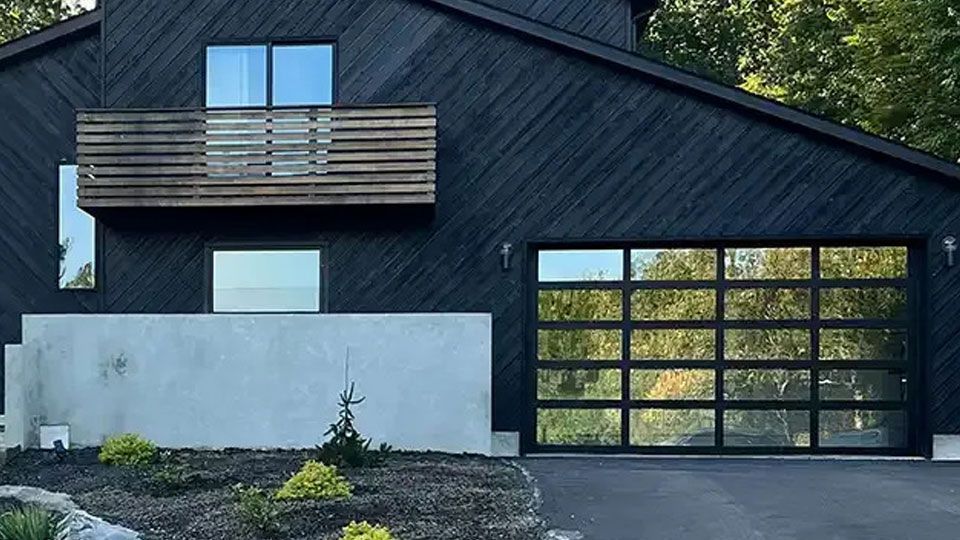 Modern dark-sided house with sloped roof, balcony, and garage surrounded by trees