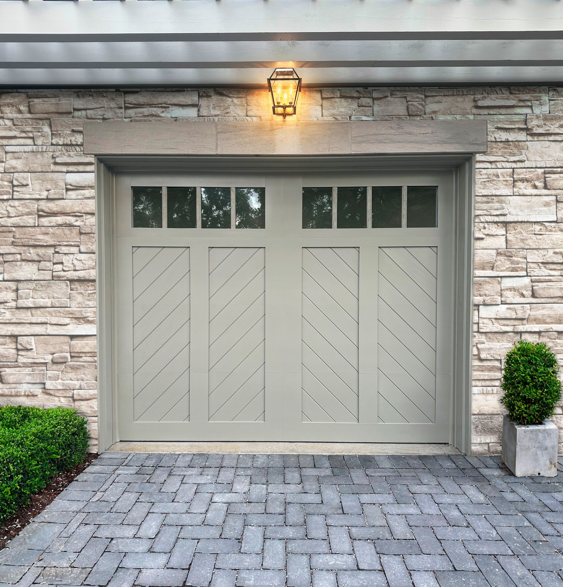 Closed light gray garage doors with diagonal panels set in a stone exterior under a porch light