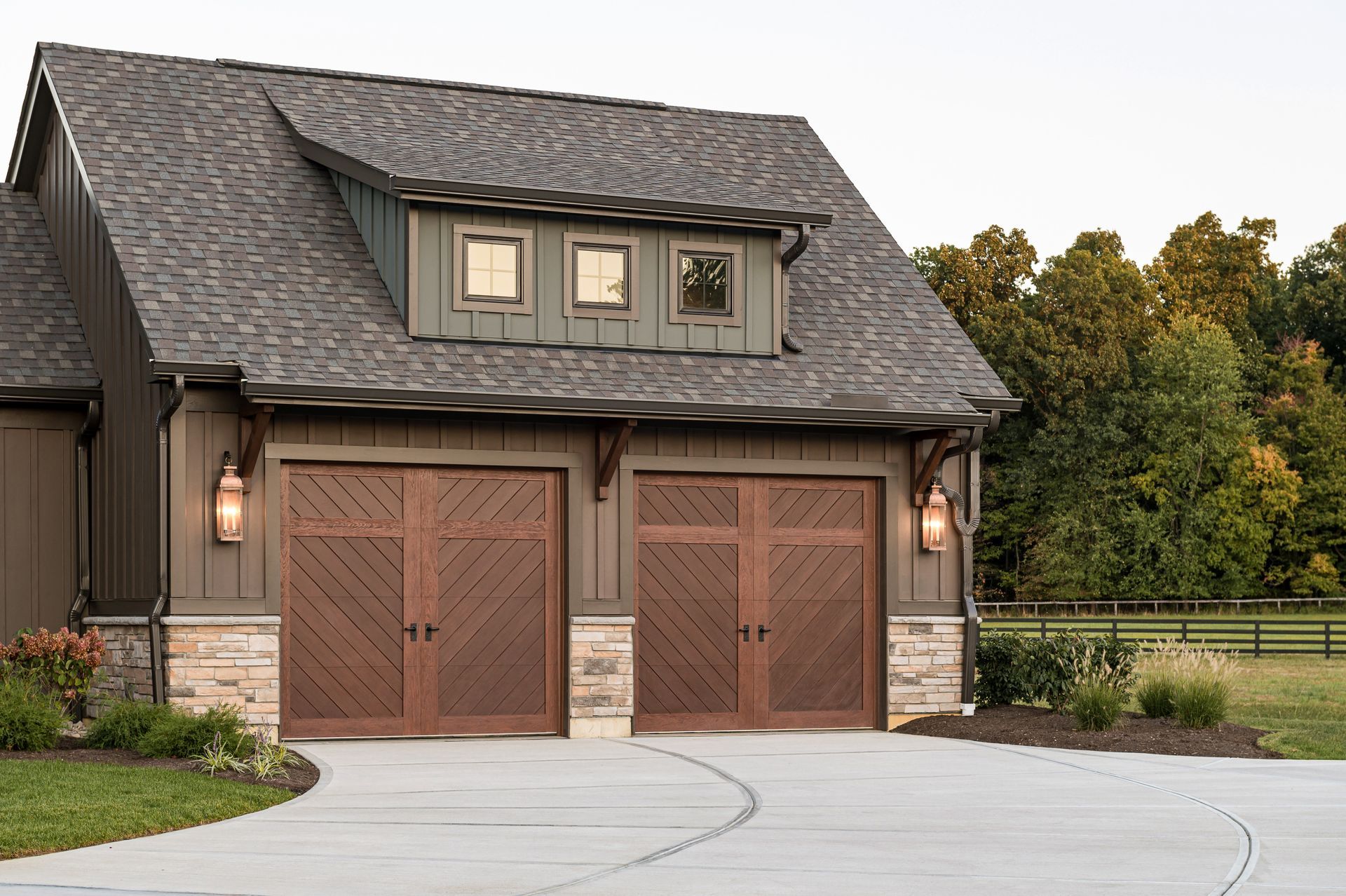 Rustic two-story garage with brown wood doors and gray shingled roof beside a curved driveway and trees