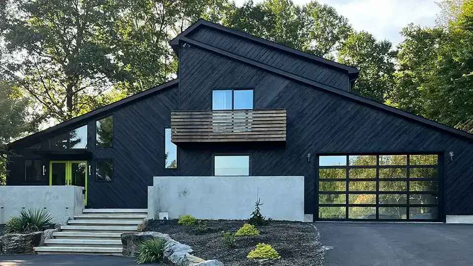 Modern dark-sided house with sloped roof, balcony, and garage surrounded by trees
