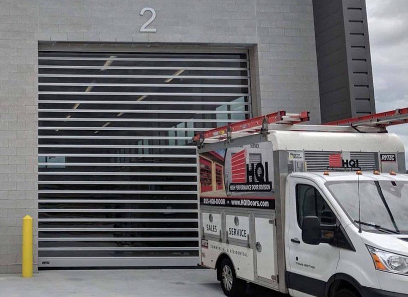 A white work van with a ladder parked in front of a horizontal-slat commercial garage door. Building number 