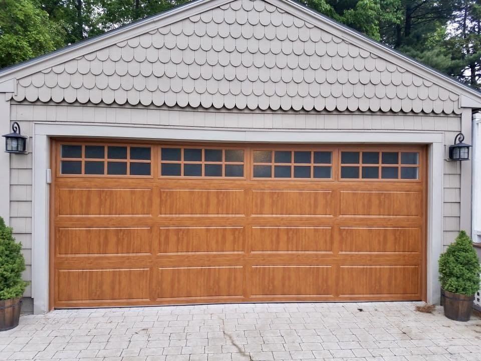 Brown garage door with windowed top, tan trim, under a gray, scalloped roof, with flanking dark sconces.