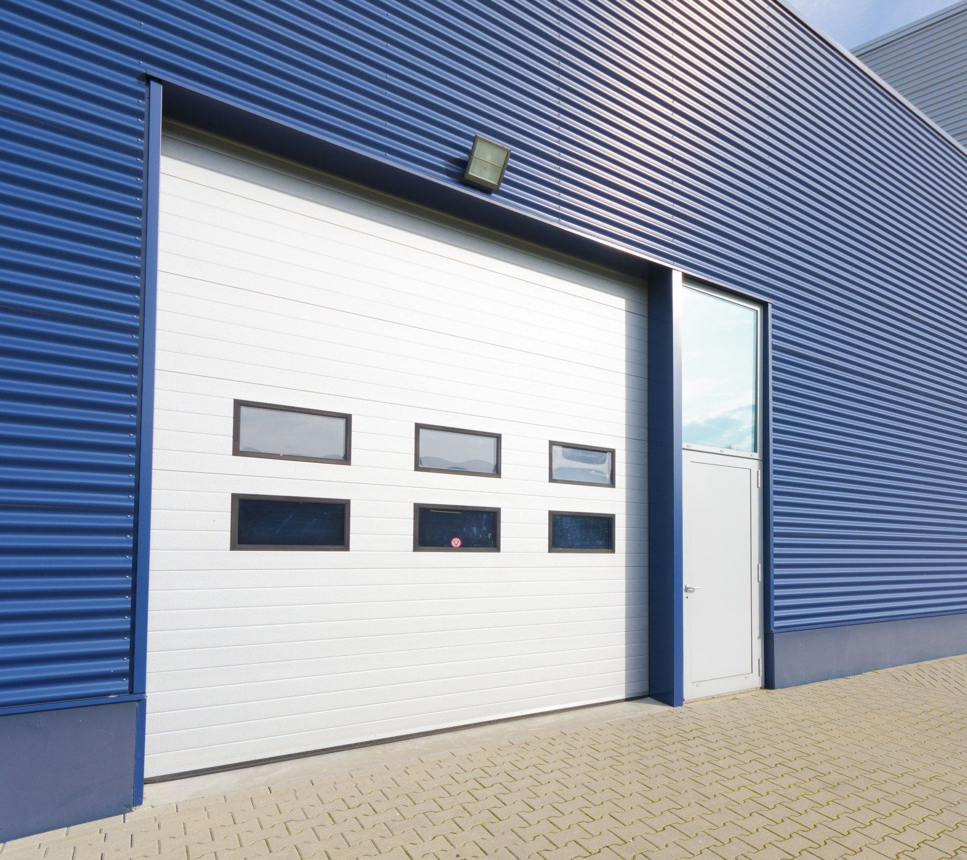 White industrial garage door with windows, on a blue building, with a side door and a brick ground.