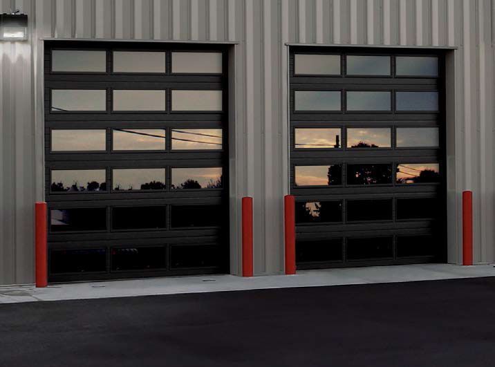 Two dark commercial garage doors with glass panels, flanked by red bollards, in a metal building.