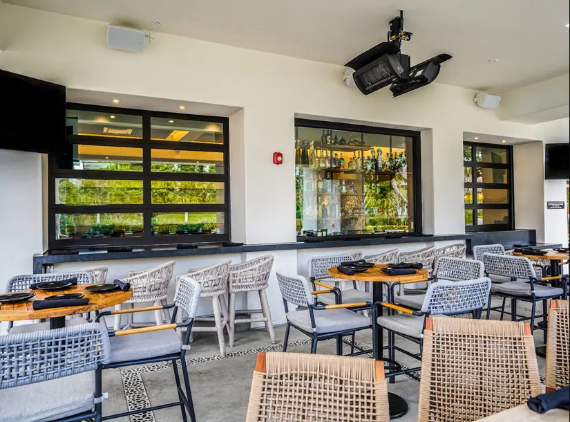 Outdoor dining area with tables, woven chairs, and bar seating along a wall with large windows, under a white stucco ceiling.