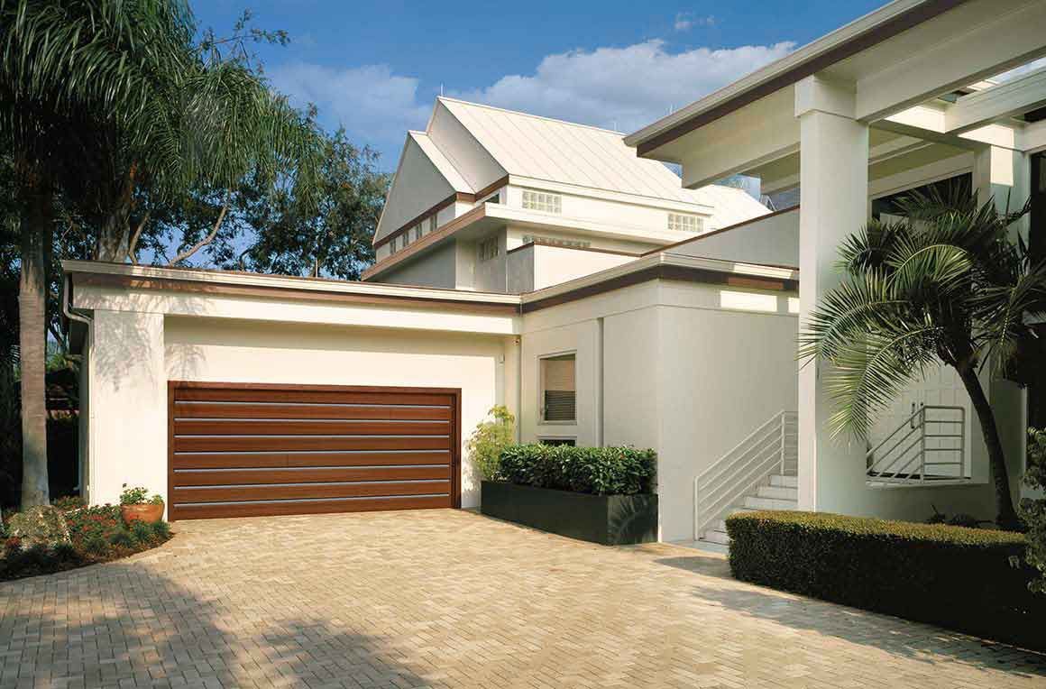 Brown garage door on a beige house with a stone driveway, palm trees and bright sunlight.