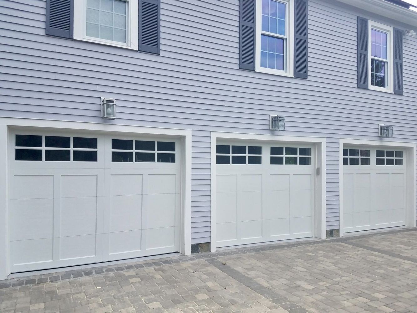 Three white garage doors with windows, gray siding, and outdoor lights.
