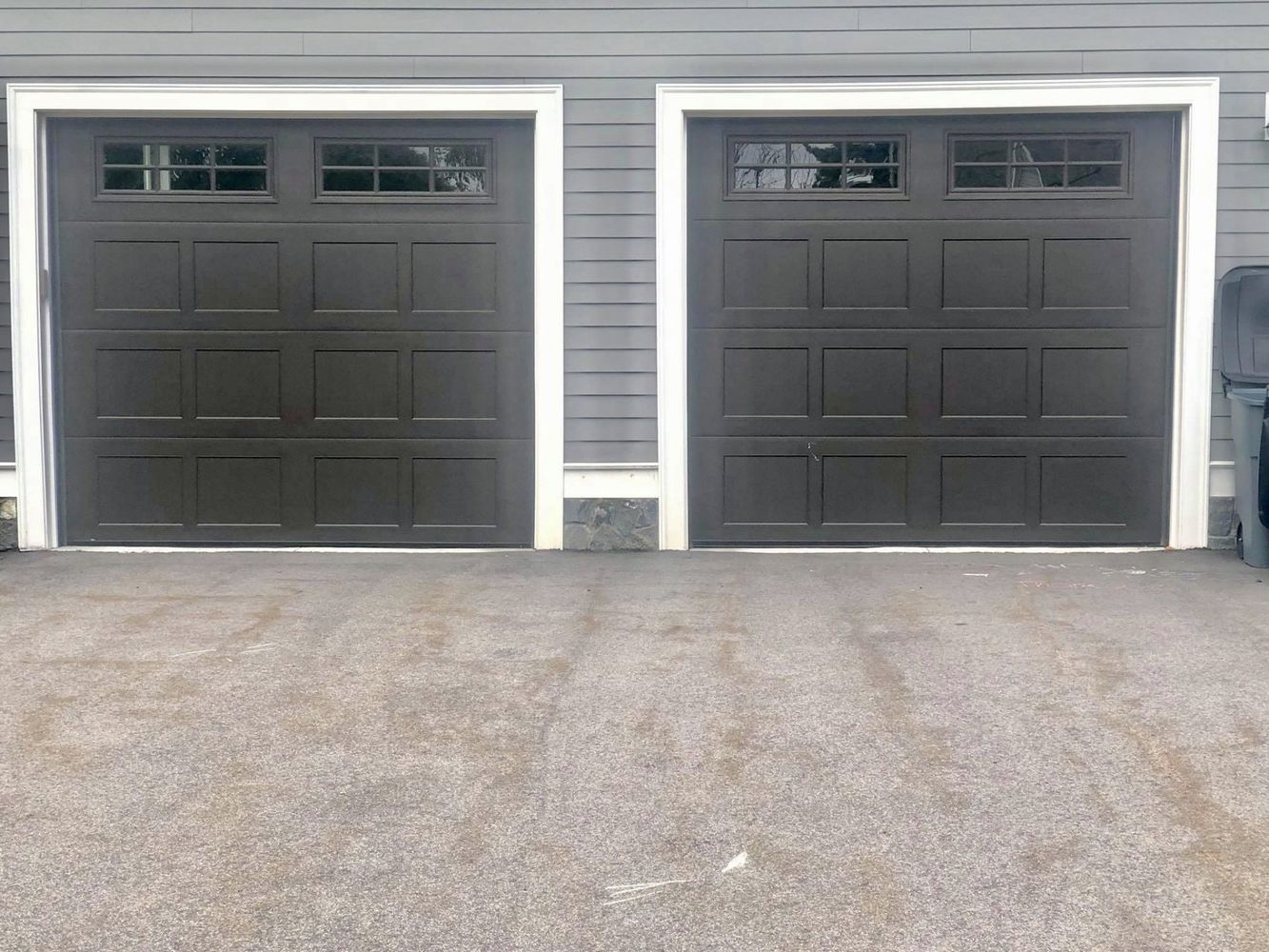 Two dark brown garage doors with white trim and windows, against a gray house.