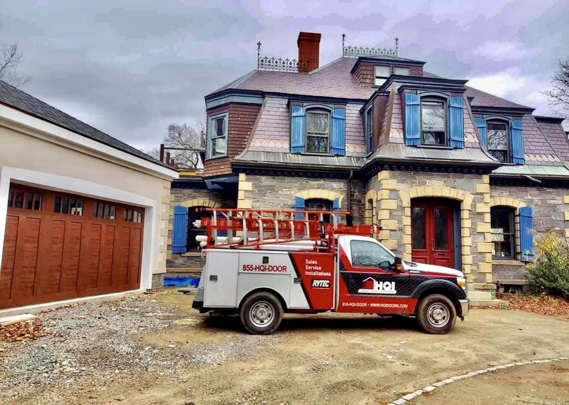 Service truck parked in front of a stone house with a garage. Brown garage door, red truck. Overcast sky.