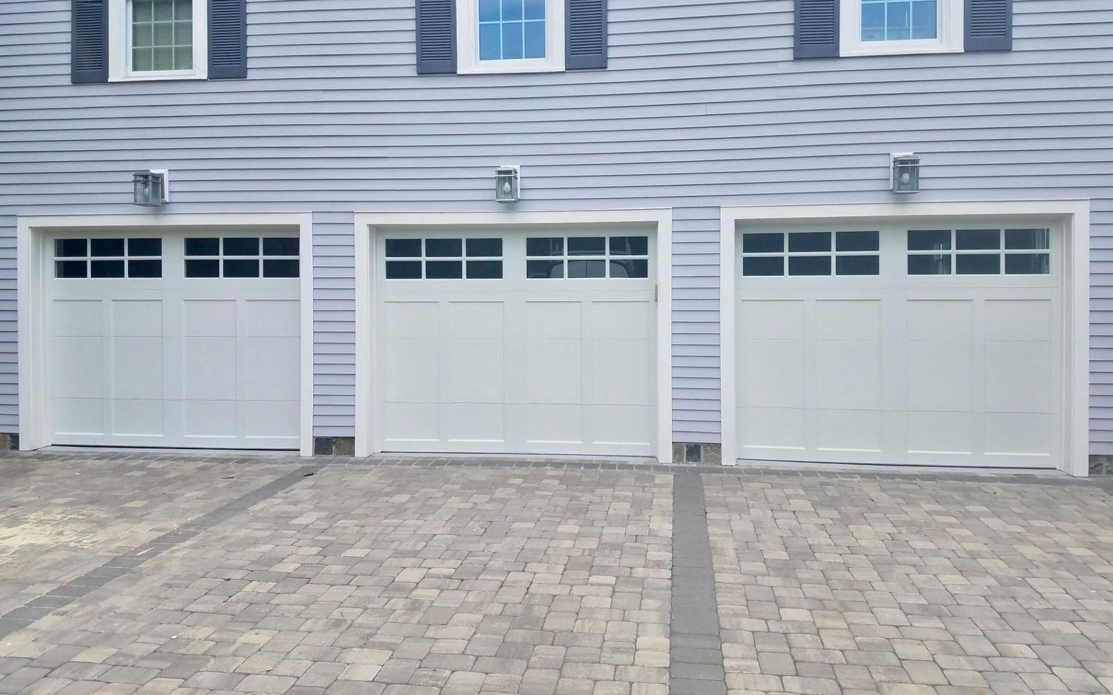 Three white garage doors with windows, mounted on a light blue building with a brick driveway.