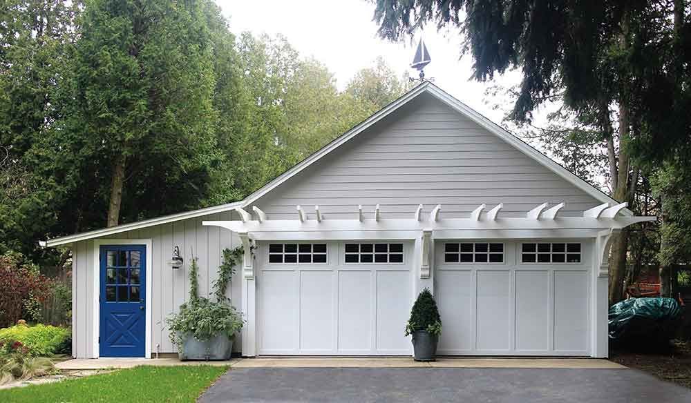 White garage with two doors and a blue door to the side, under a pergola, set among trees.