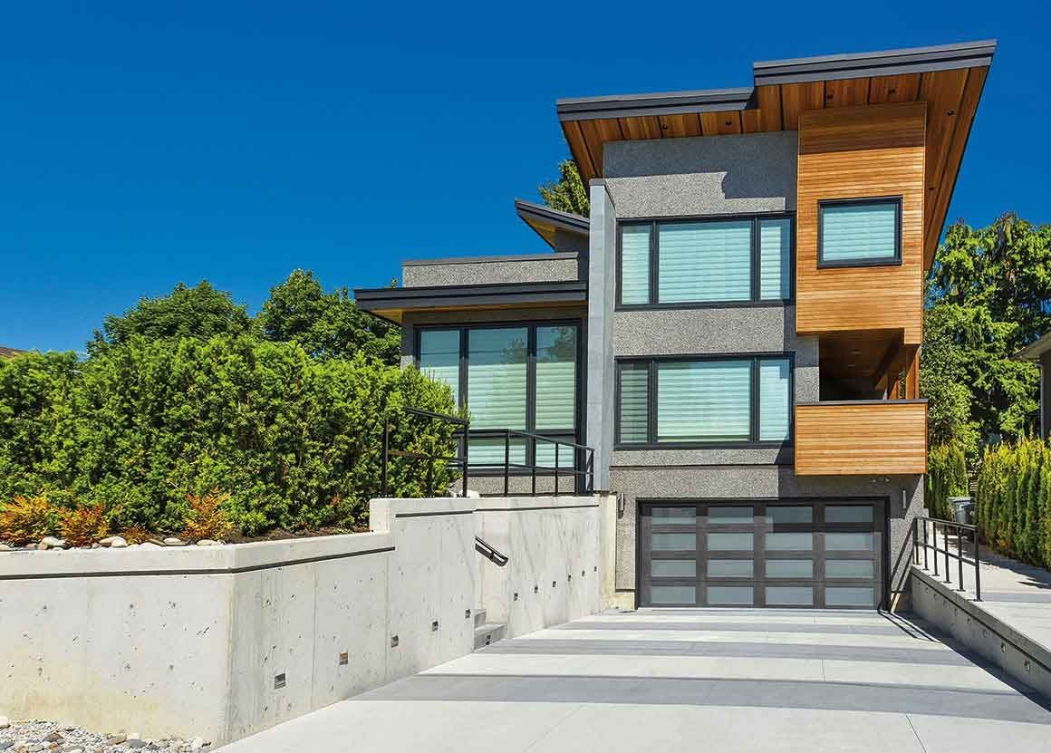 Modern home with gray stucco, wood accents, and garage. Concrete retaining wall and blue sky.