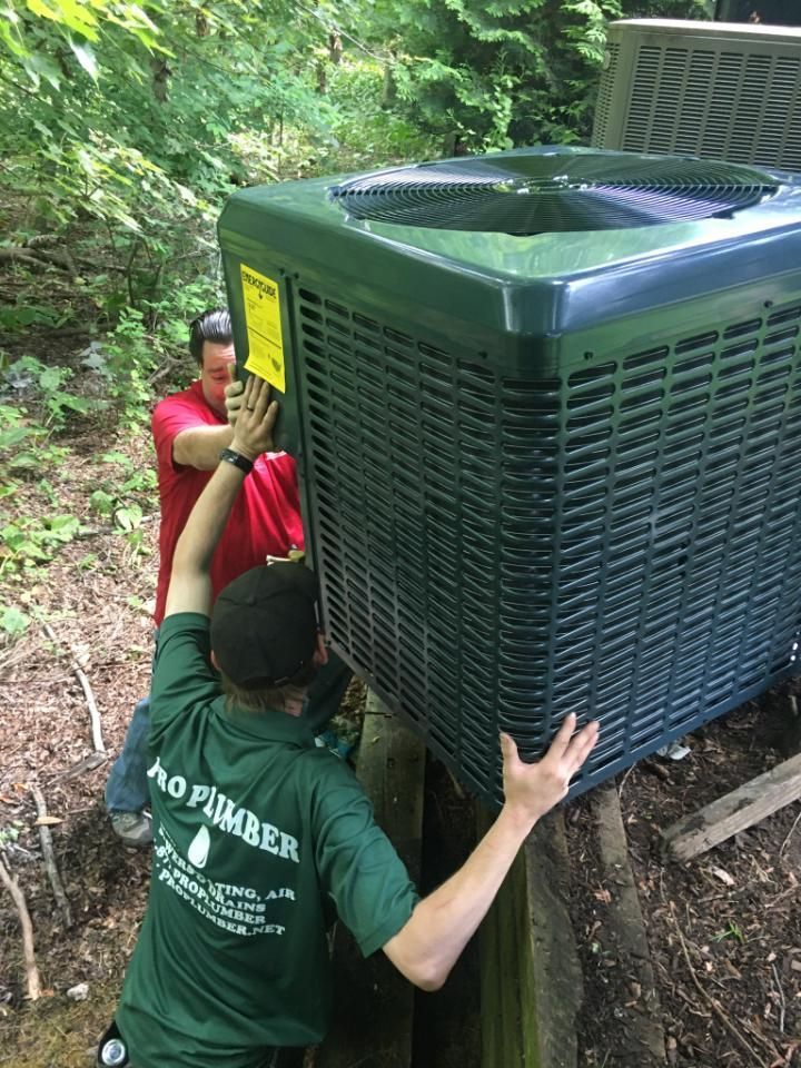 Two people maneuver a large outdoor HVAC unit through a wooded area.