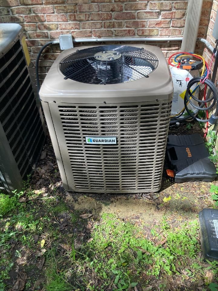 Outdoor air conditioner unit with fan, sitting beside brick wall and yard equipment.