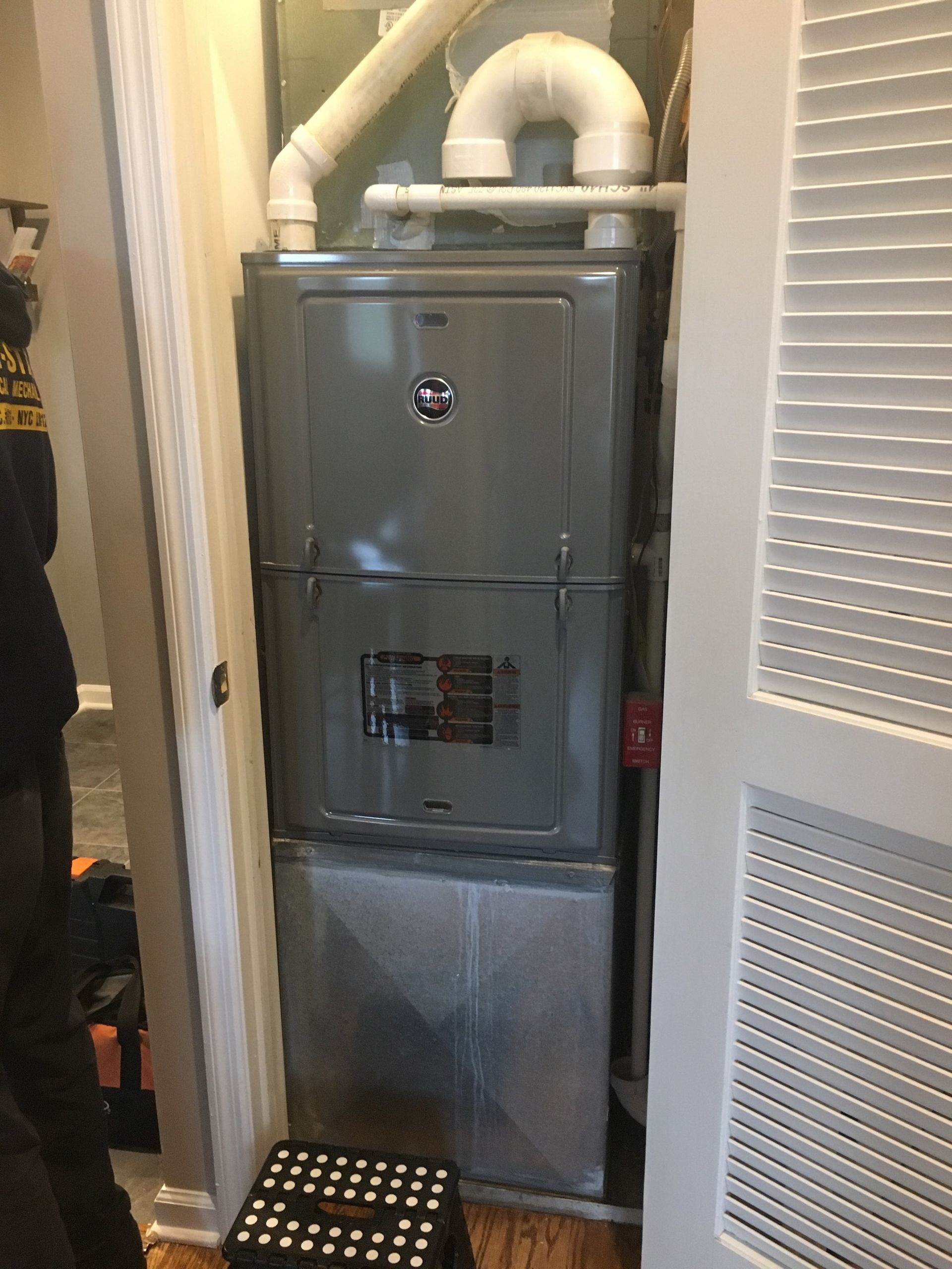 Gray furnace in a narrow utility closet with white ductwork above and louvered door on the right