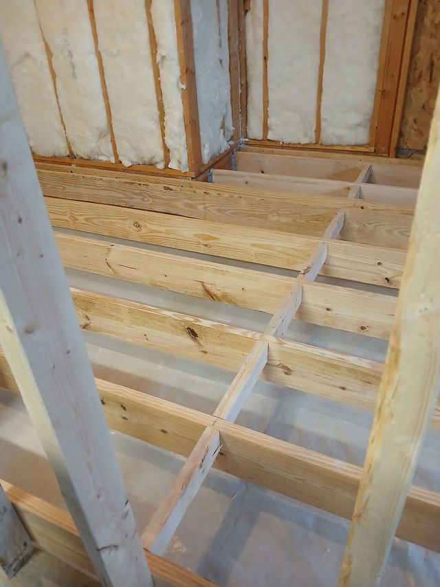 Framed wooden floor joists in a house under construction with exposed insulation and wall studs.