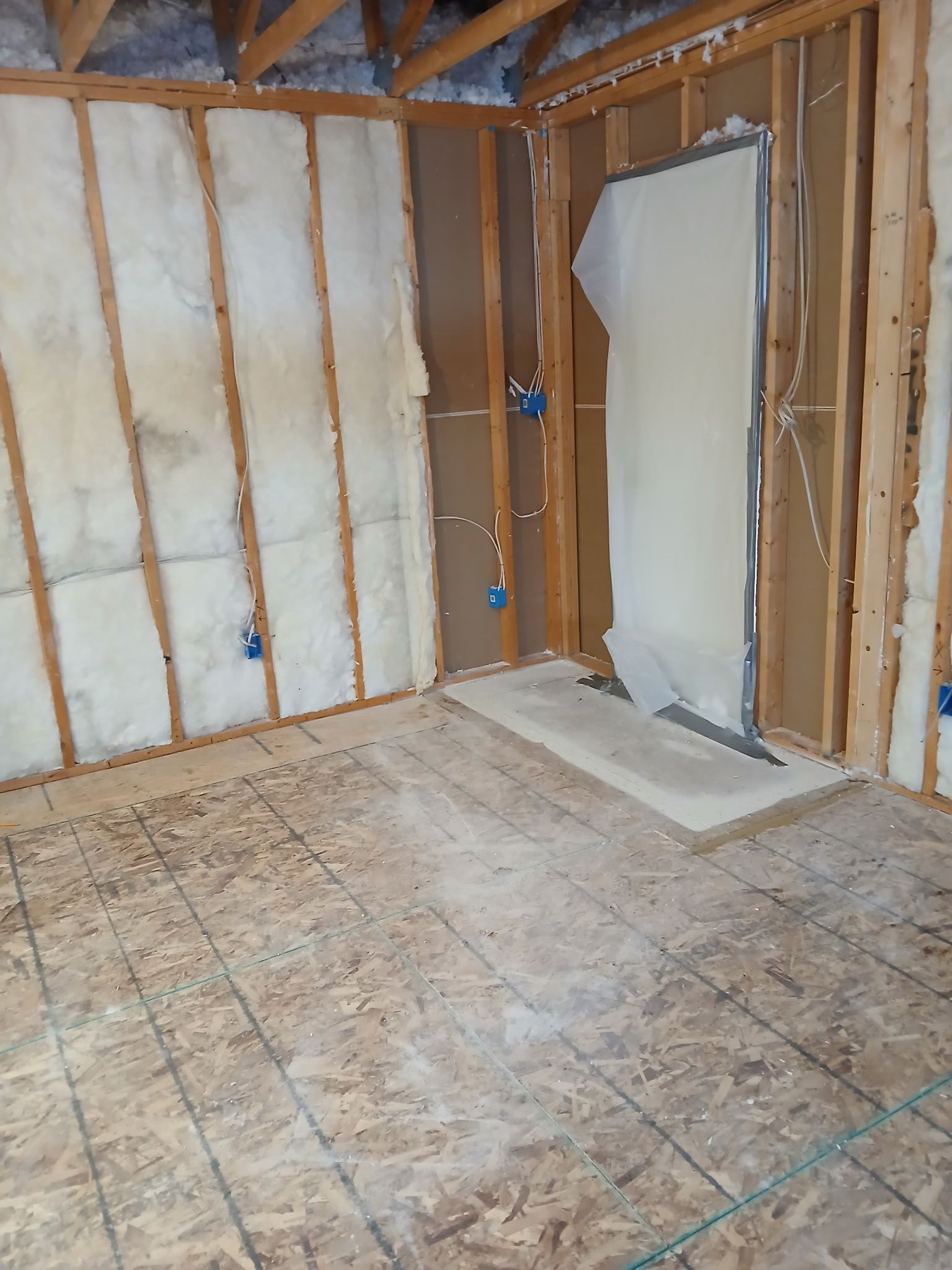 Partially renovated room with exposed studs, insulation, plywood floor, and a doorway opening to another room