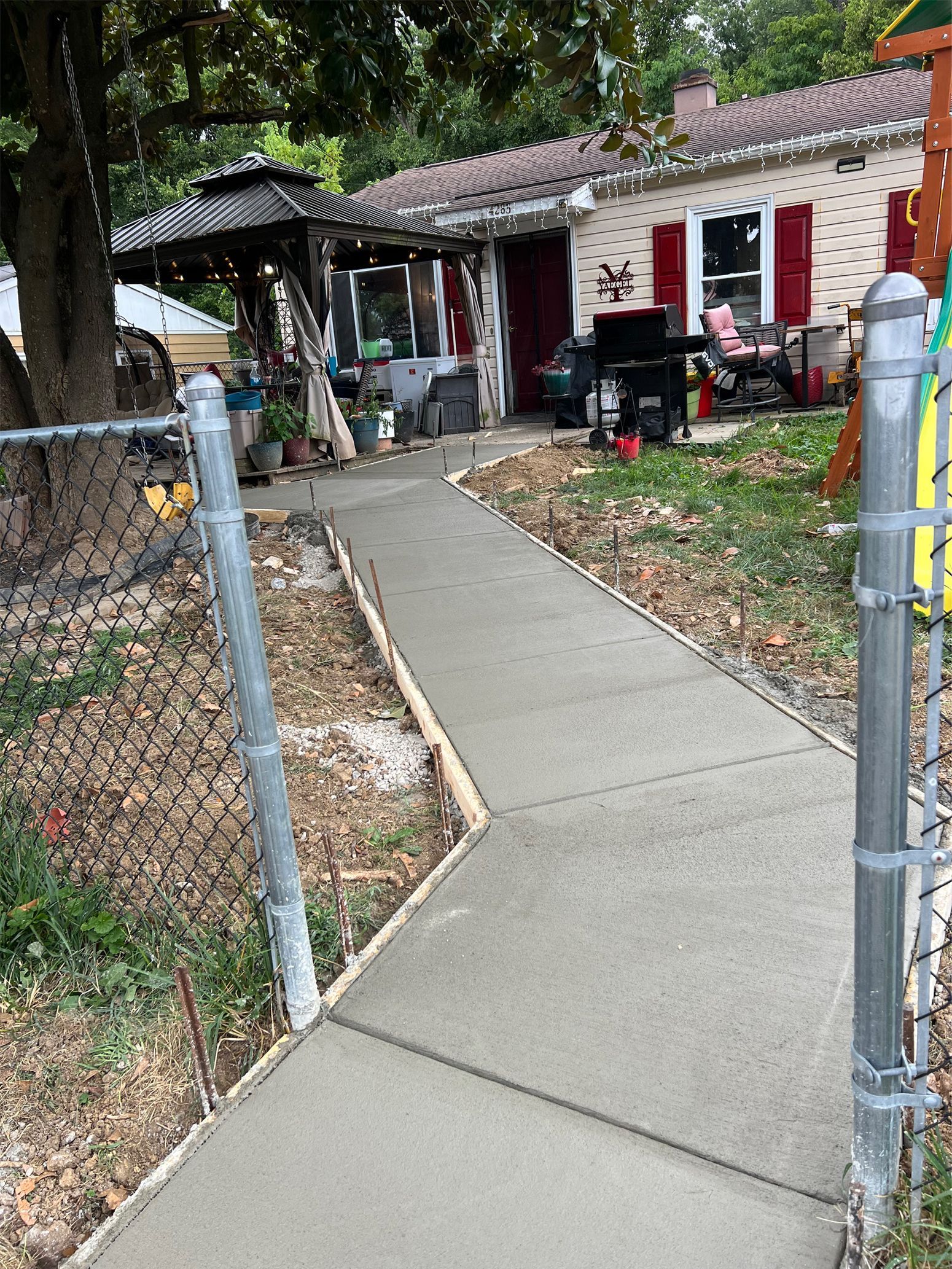 Concrete walkway leading to a red house with parked scooters and a chain-link fence in a leafy yard