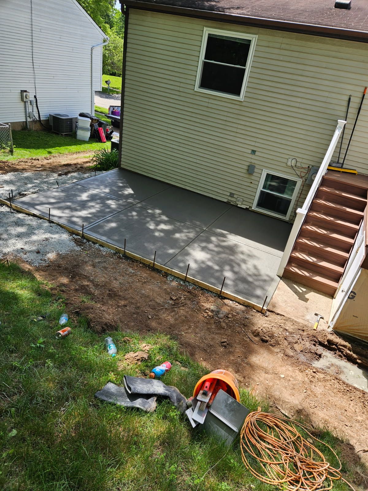 Backyard concrete patio beside beige house with stairs and small windows; yard debris in foreground