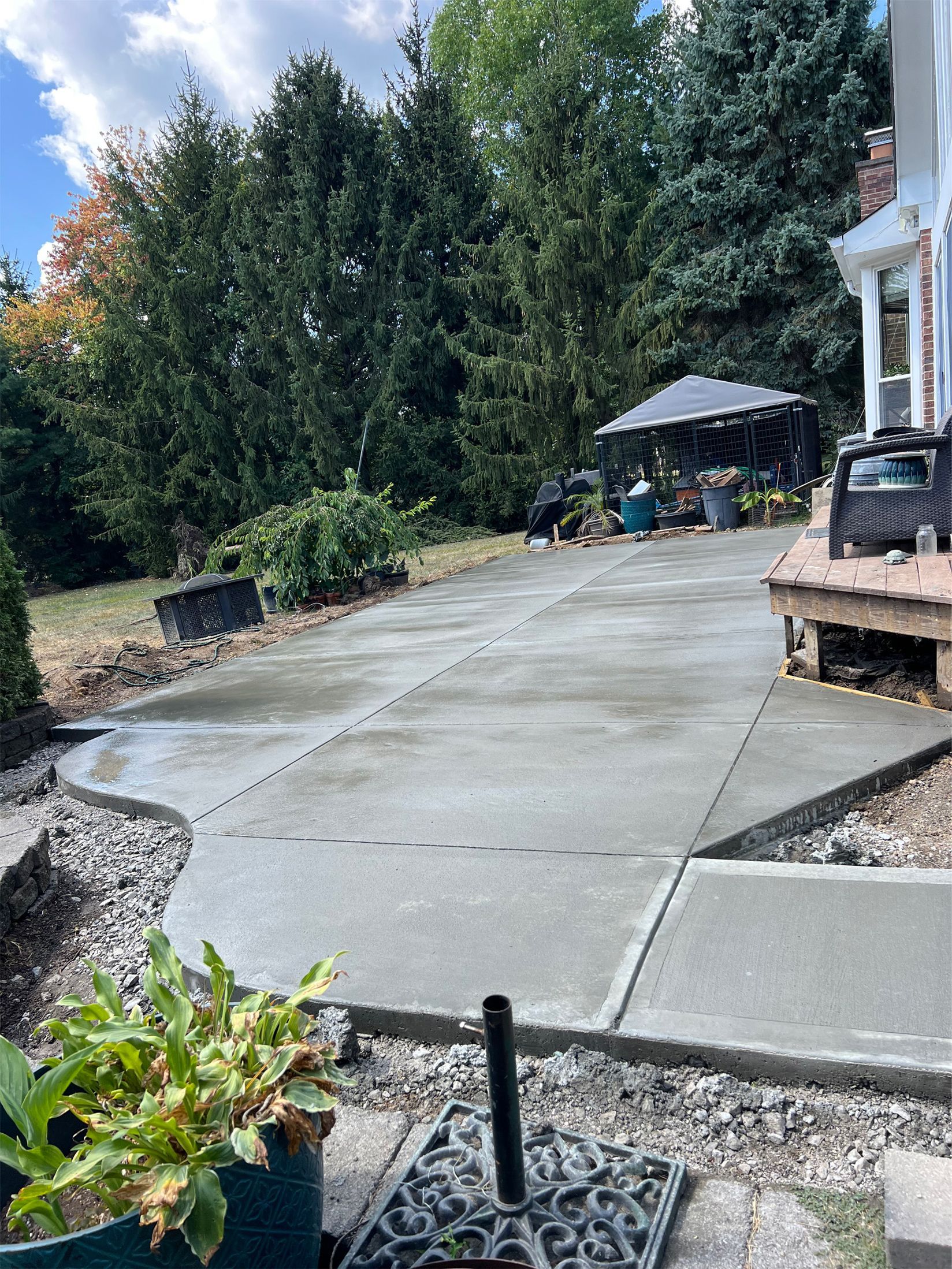 Concrete patio and walkway beside a house, with trees, shrubs, and a patio umbrella in the background