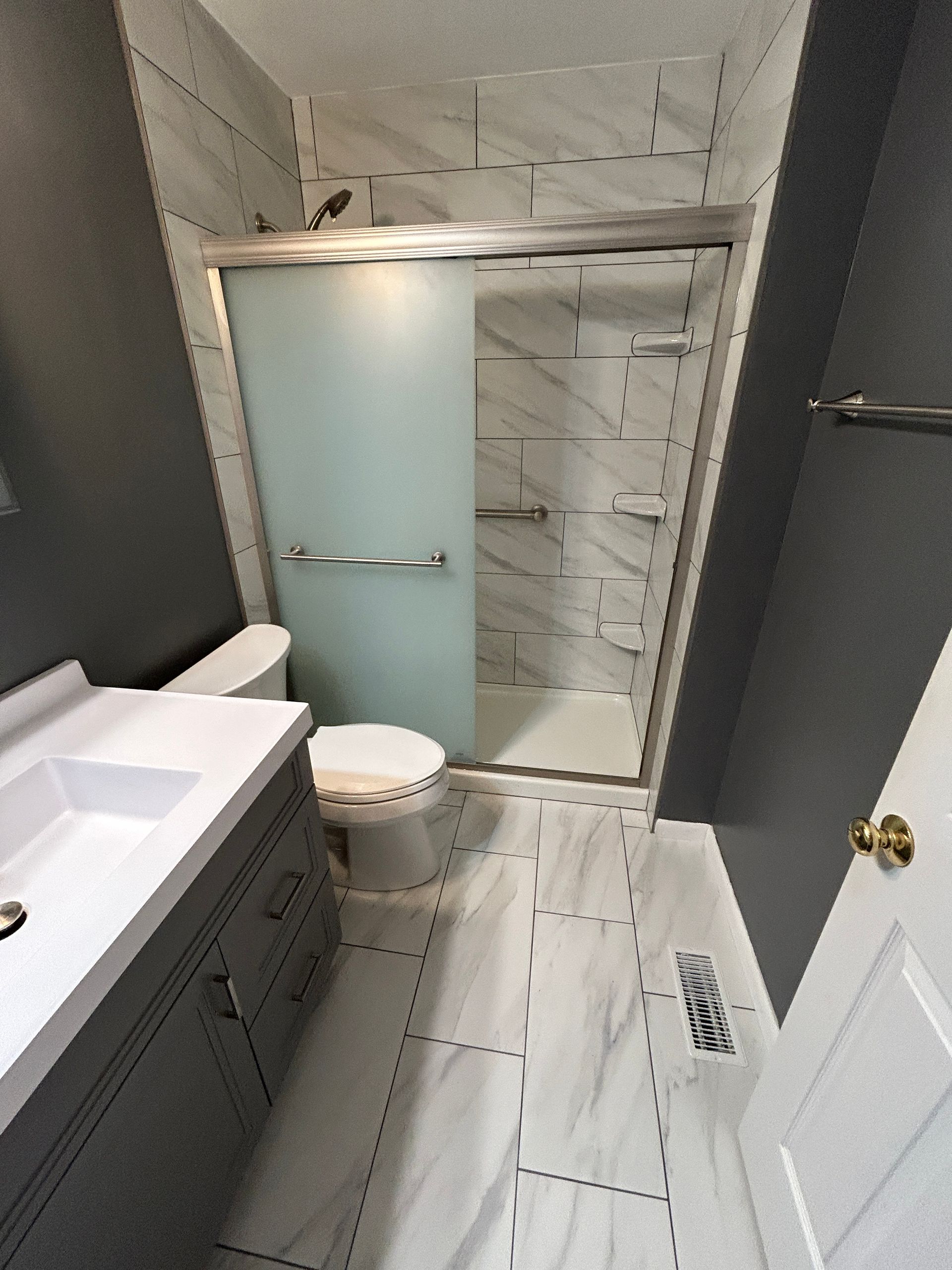 Small bathroom with white tiled shower, toilet, vanity, and gray door.