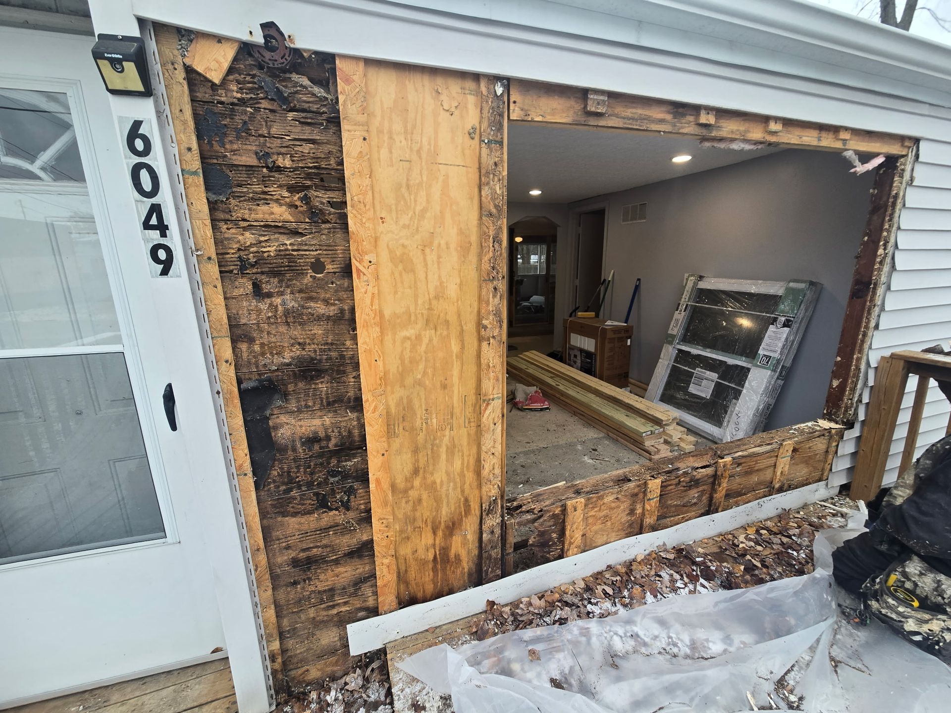 Damaged front wall of a house with exposed framing, plywood patch, and broken window opening above snowy steps