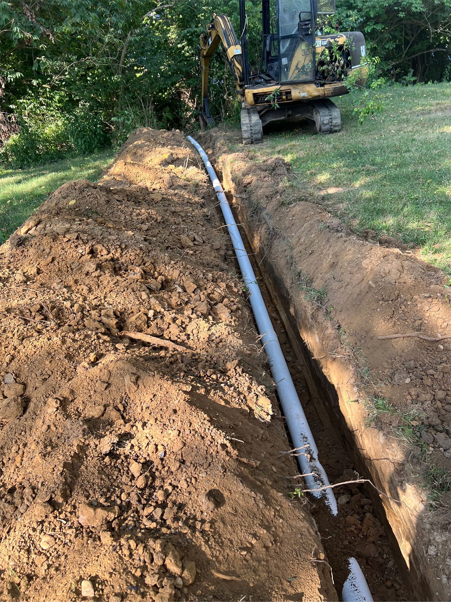 Excavator trenching a yard for a buried black pipe beside a dirt mound and trees