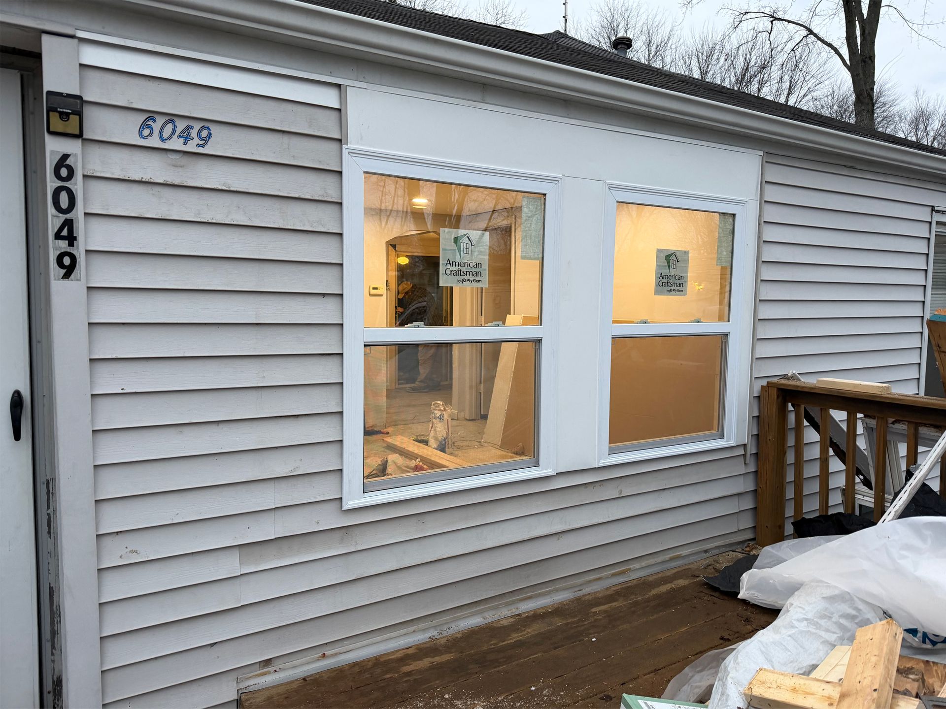 White-sided house exterior with a lit window and number 6049 beside a wooden deck in winter.