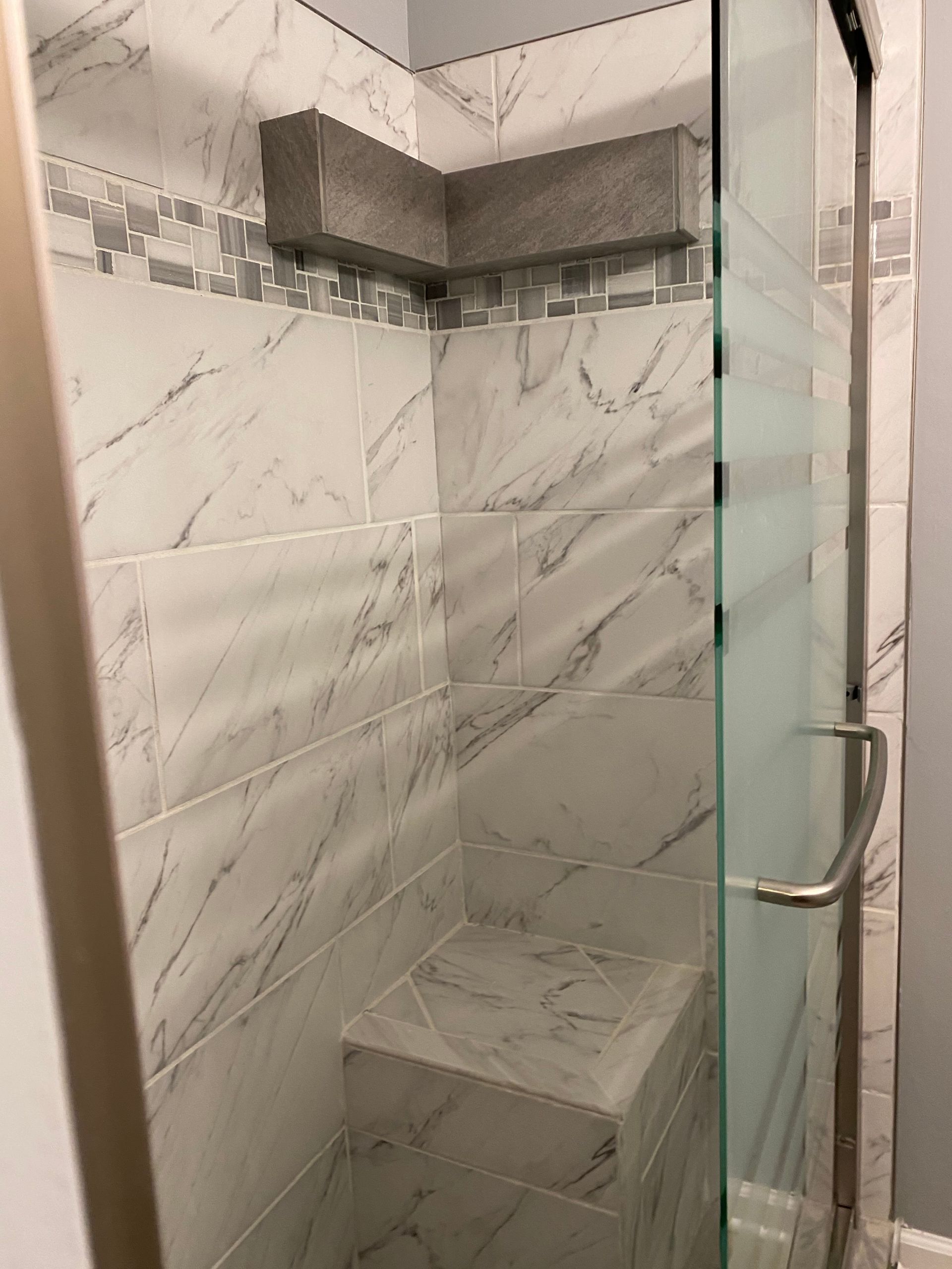 Tiled shower stall with glass door, built-in bench, and gray marble-pattern walls