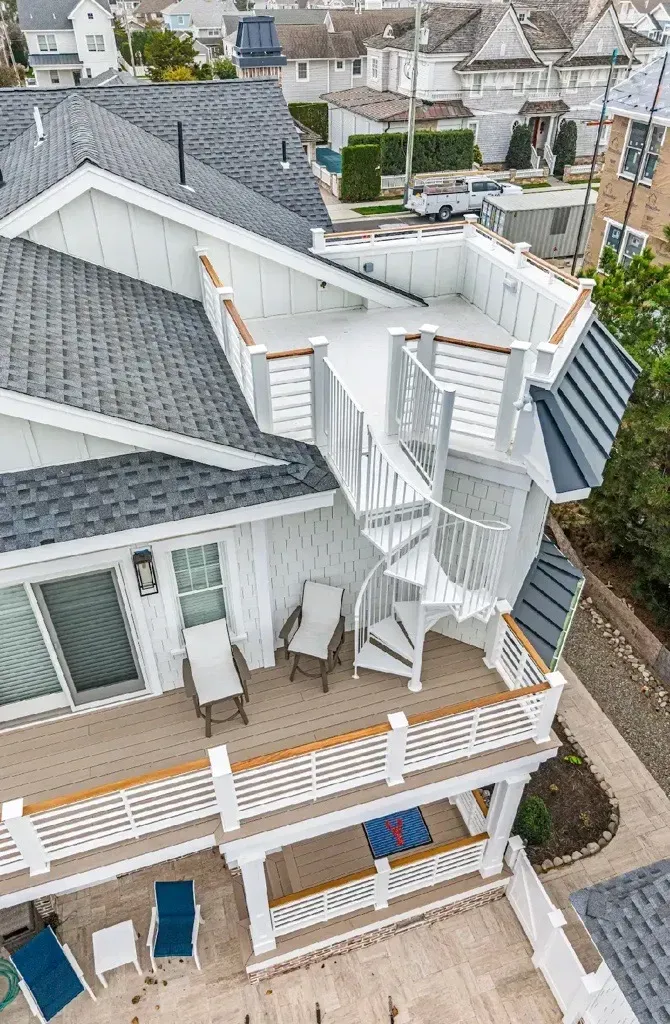 A high-angle view of a white multi-level house exterior with decks, a spiral staircase, and gray shingled roof sections.
