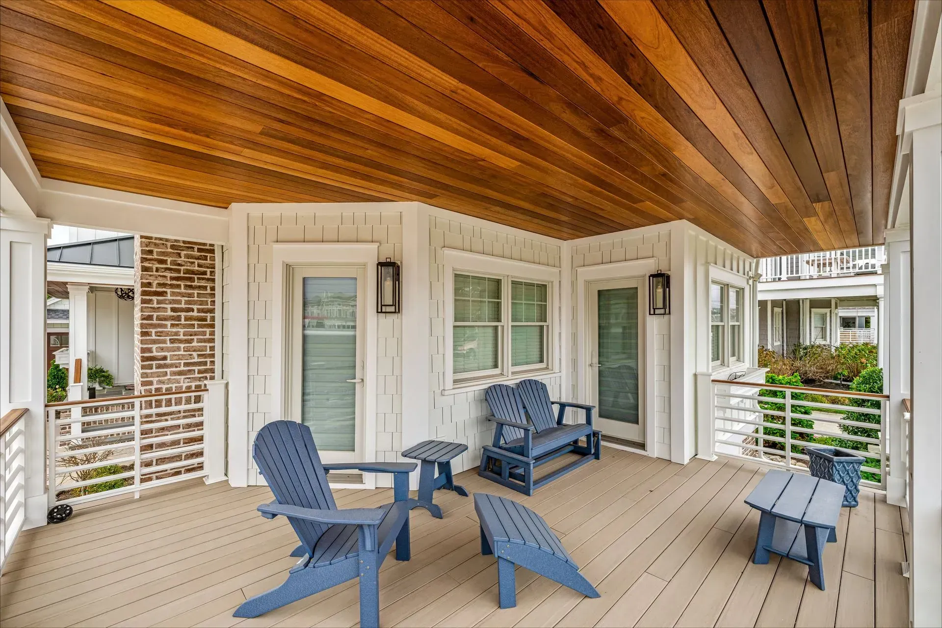 Covered porch featuring blue Adirondack chairs, a small table, and a wooden ceiling on a white home with brick accents.