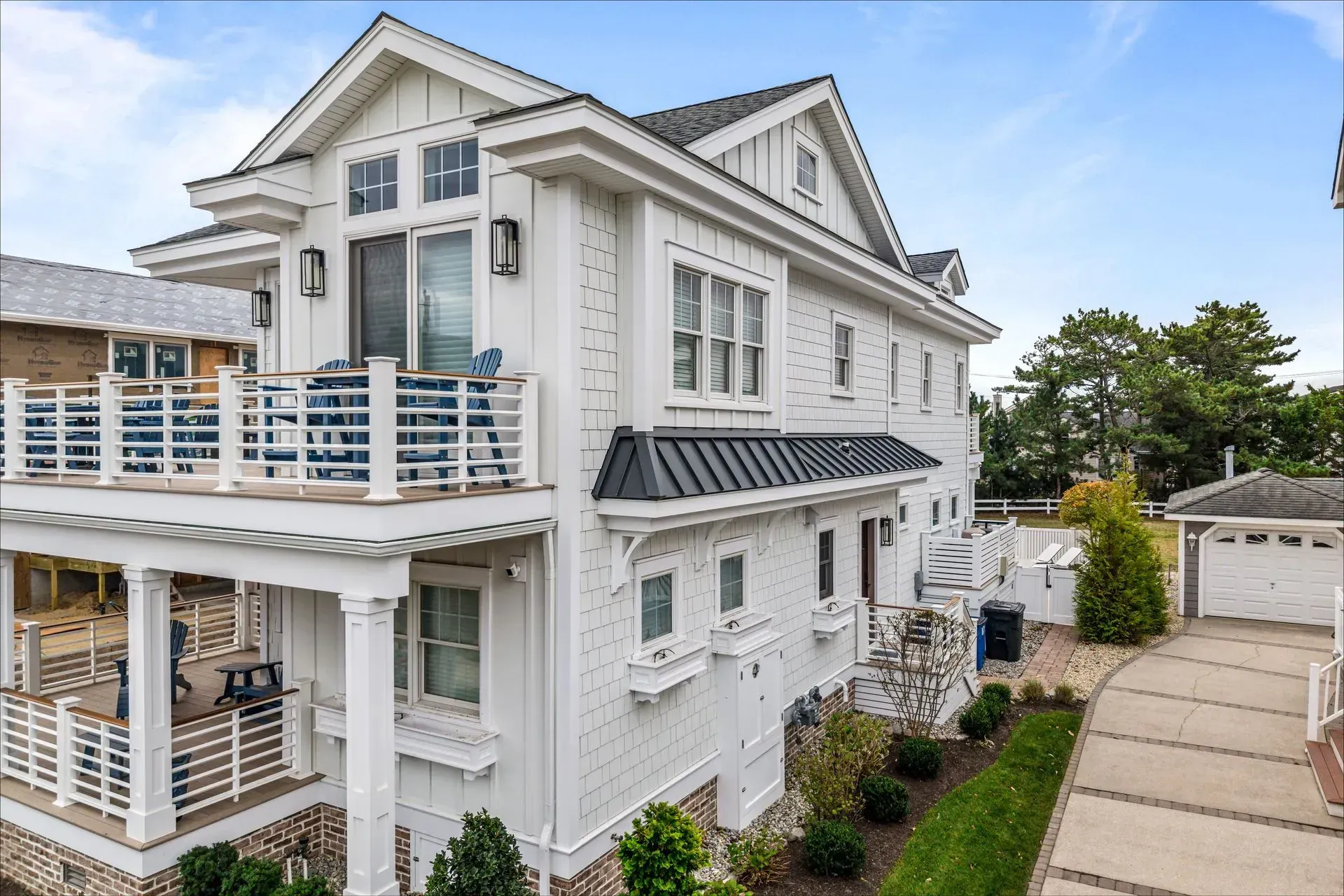 A modern, two-story white coastal house with a covered patio, balcony, dark roof, and a paved driveway.