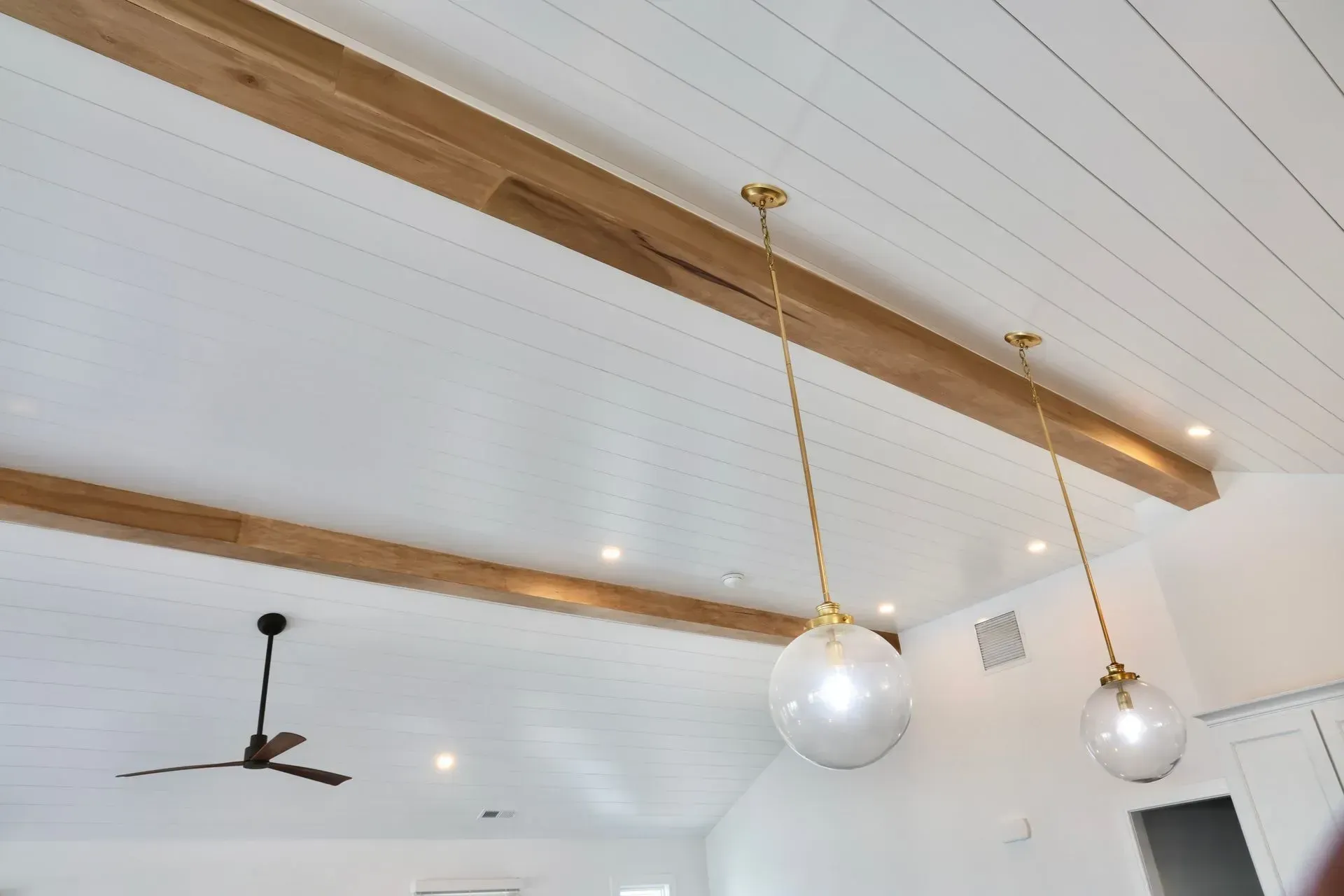 A white paneled, vaulted ceiling features two wooden beams, brass pendant lights, and a ceiling fan.