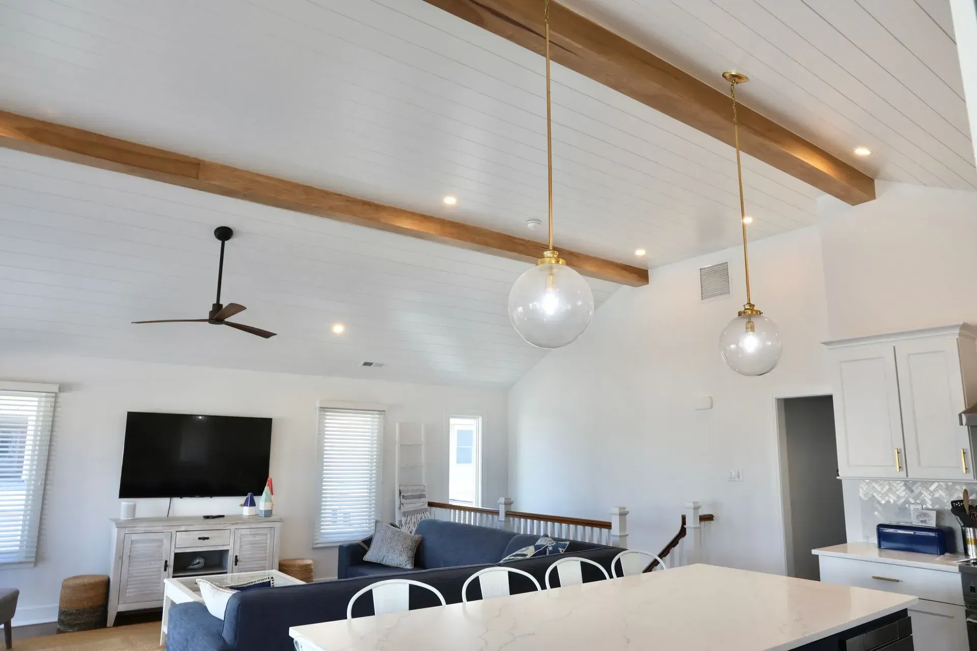Modern open-concept living area with white walls, a vaulted shiplap ceiling, wood beams, pendant lights, and a blue sofa.