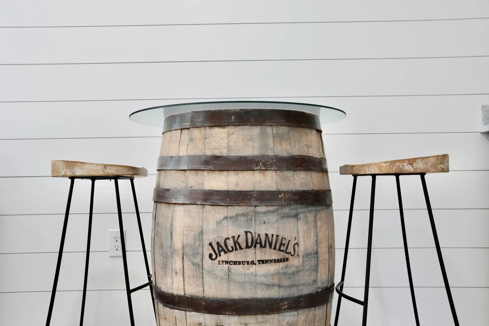 A Jack Daniel's whiskey barrel converted into a pub table with a glass top, flanked by two rustic wooden bar stools.