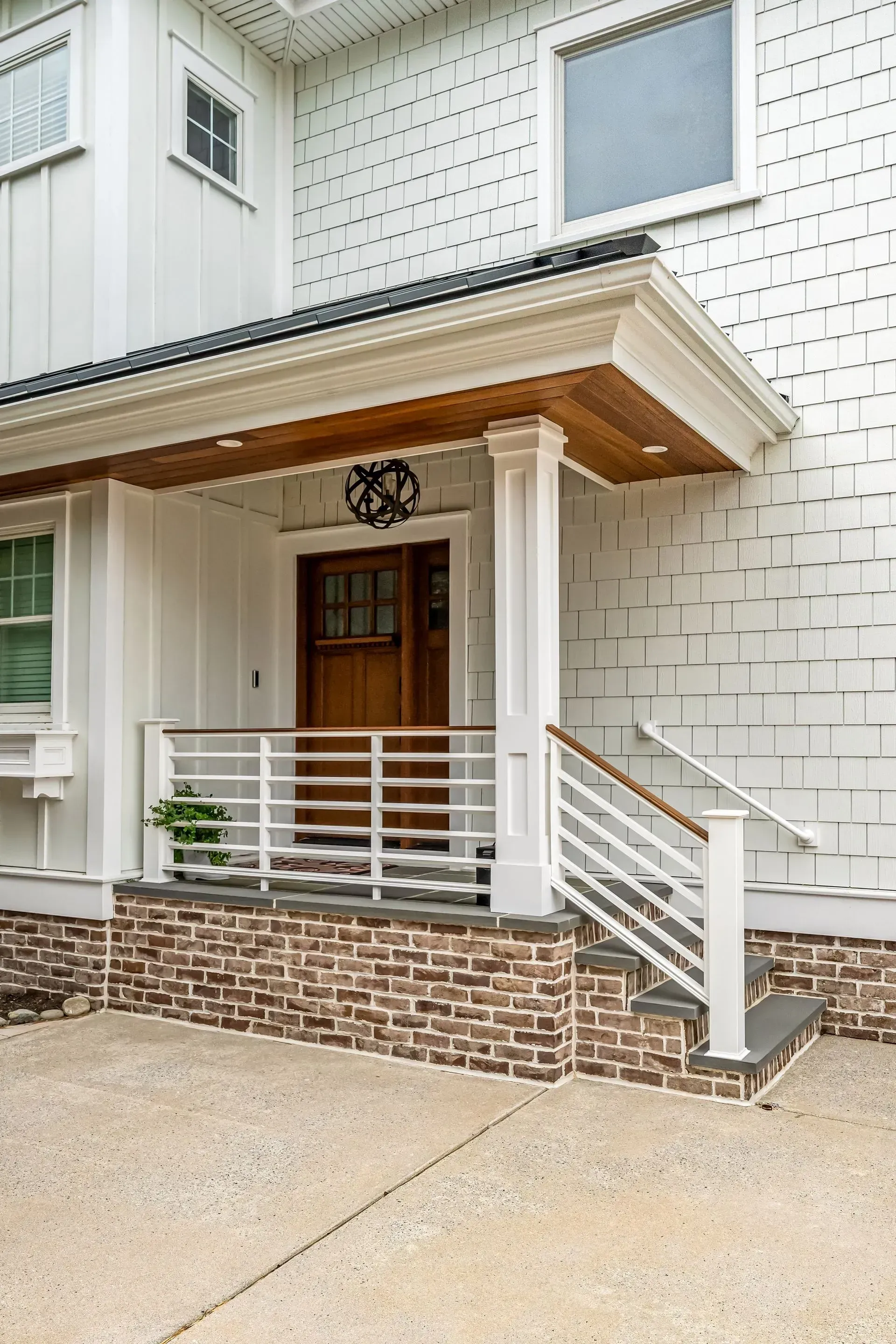 The front entrance of a modern white house features a wooden door, a brick foundation, and a white metal porch railing.