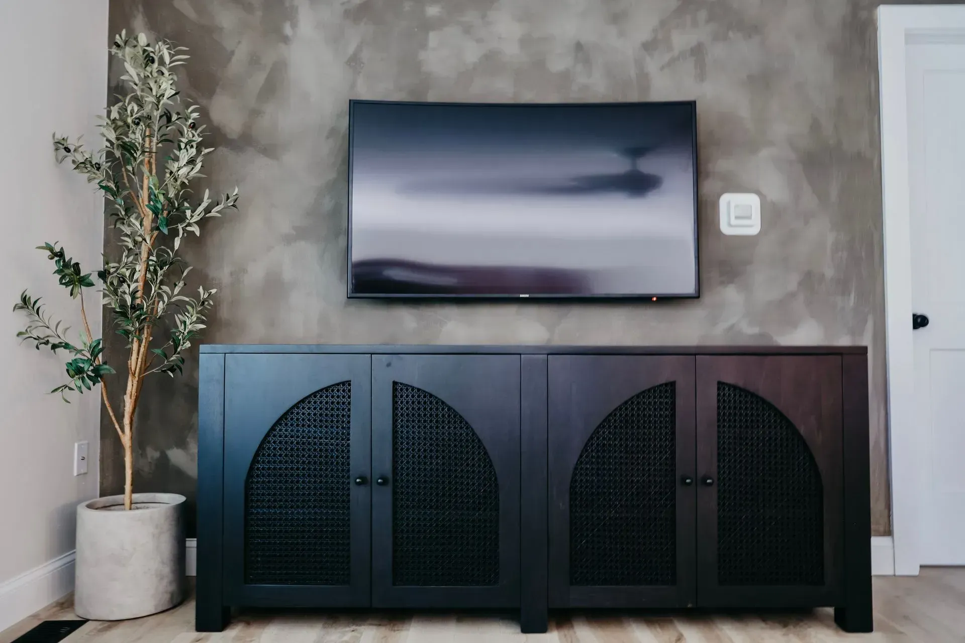 A dark cabinet with arched, latticed doors sits against a gray, textured wall beneath a mounted television and a plant.