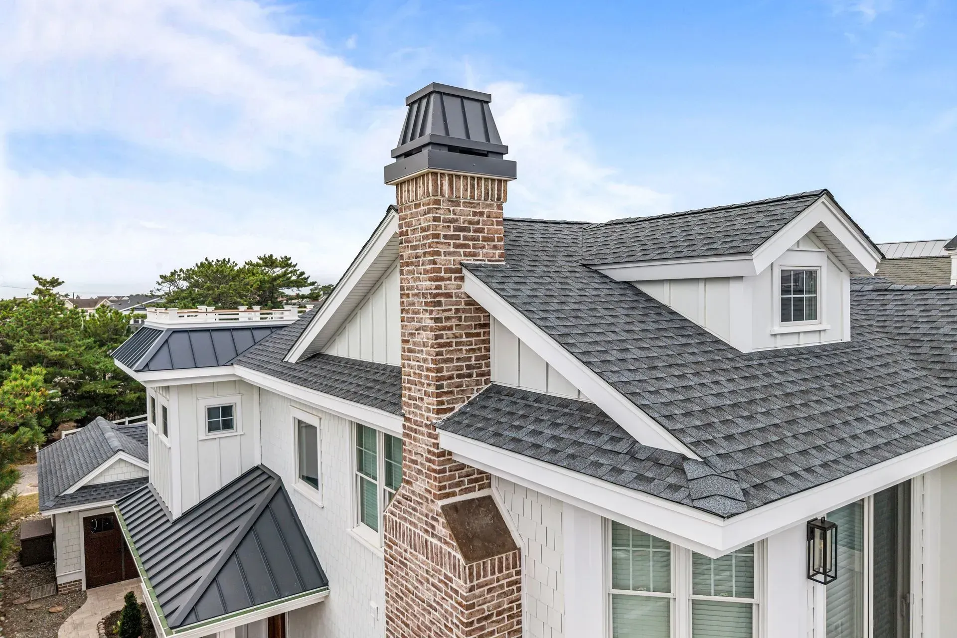 Exterior view of a house with a brick chimney, gabled roofs, white siding, and a dark metal roof accent.